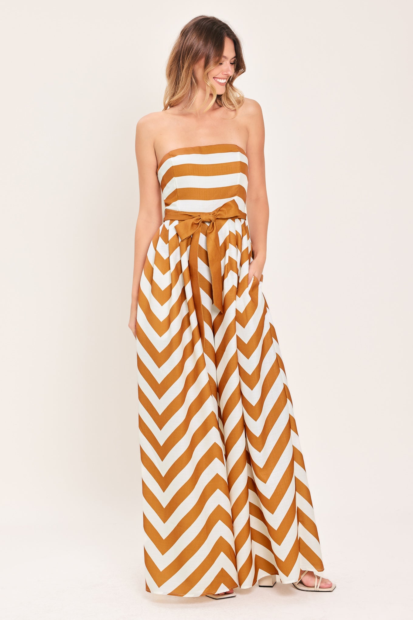 Woman wearing a camel and white strapless, long chevron-patterned jumpsuit featuring solid tie, wide leg and smocked back bodice | Flying Tomato