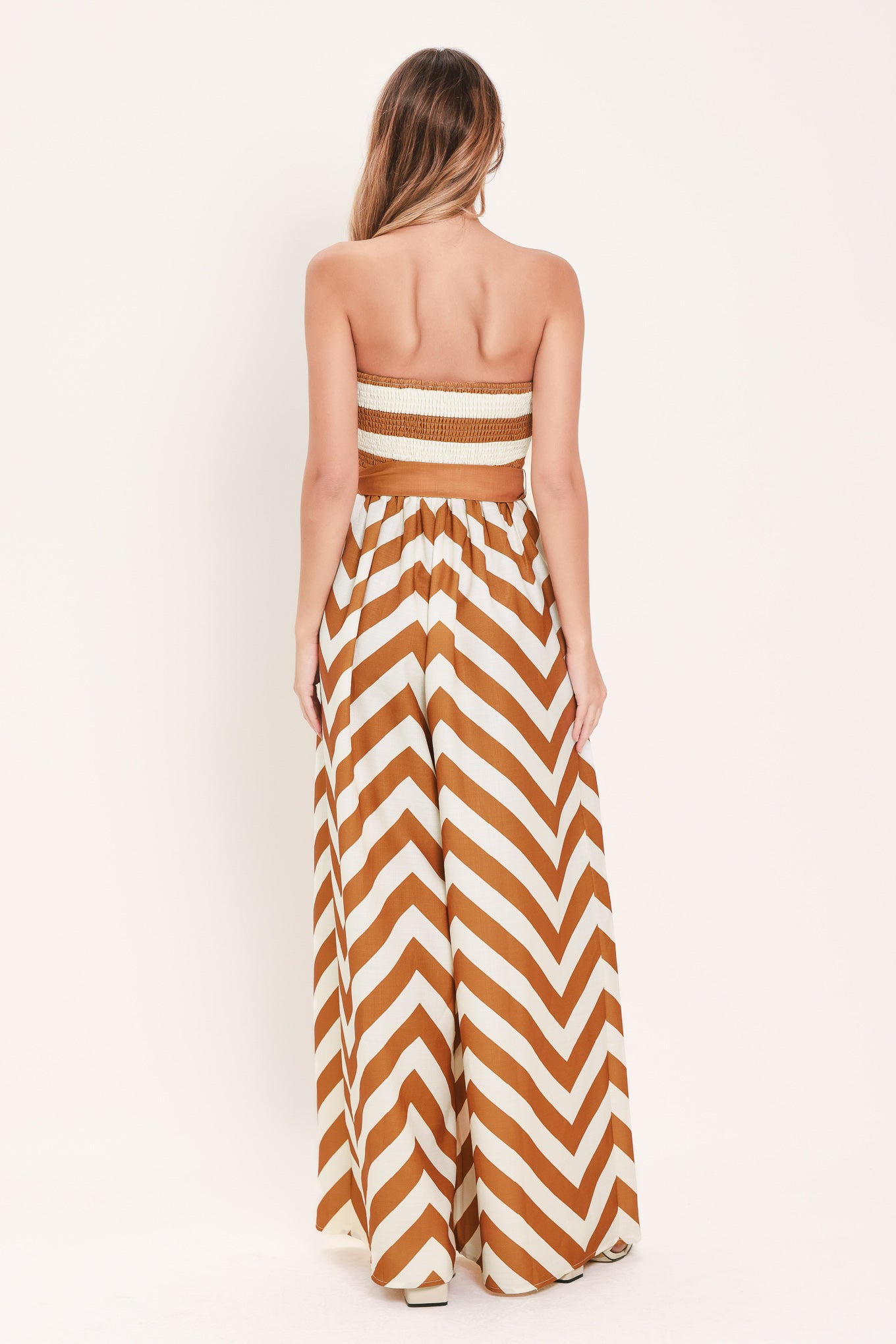 Woman wearing a camel and white strapless, long chevron-patterned jumpsuit featuring solid tie, wide leg and smocked back bodice | Flying Tomato