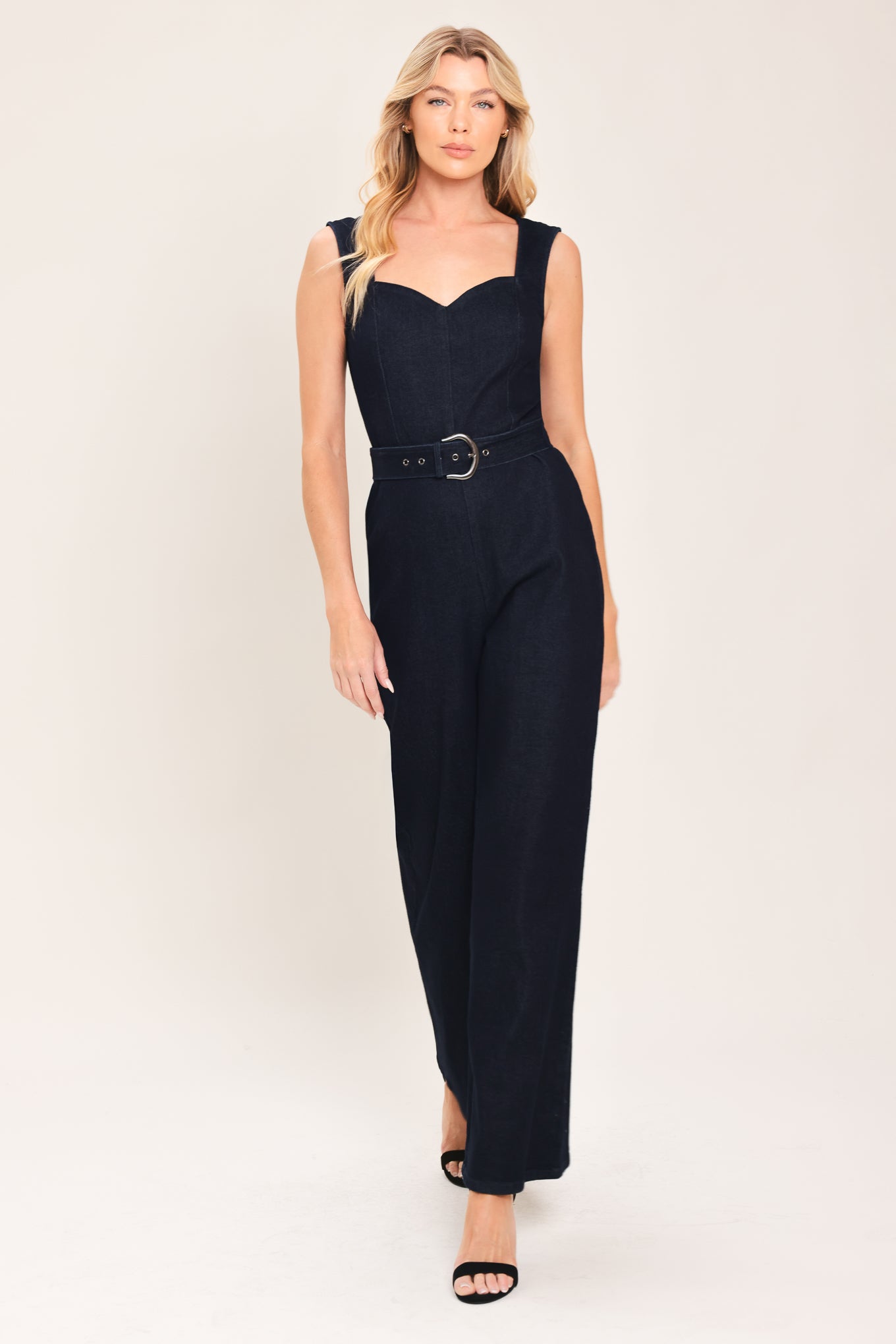Woman wearing a dark indigo denim jumpsuit featuring sweetheart neckline, sleeveless, self belt, wide leg and back zipper closure | Flying Tomato
