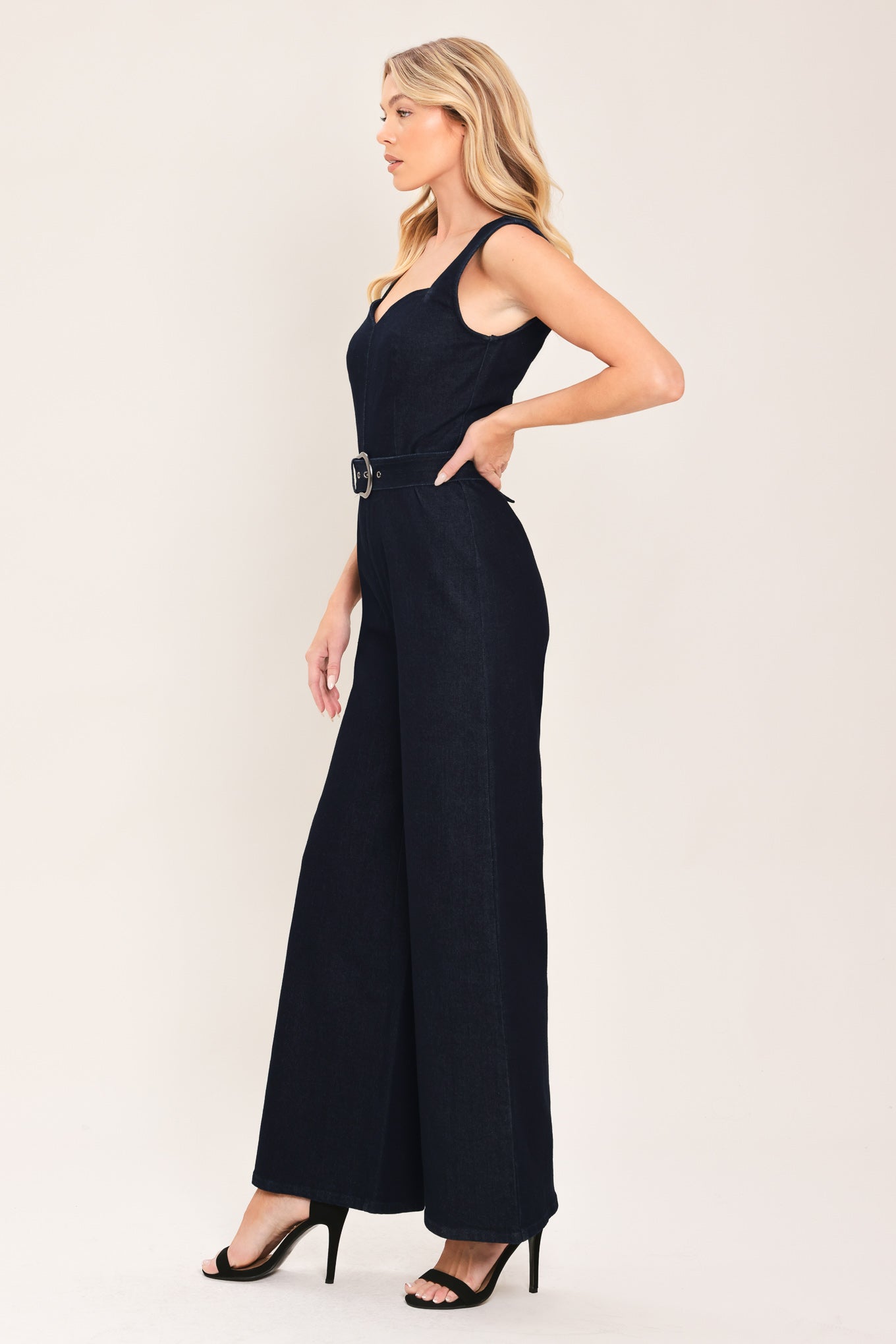 Woman wearing a dark indigo denim jumpsuit featuring sweetheart neckline, sleeveless, self belt, wide leg and back zipper closure | Flying Tomato