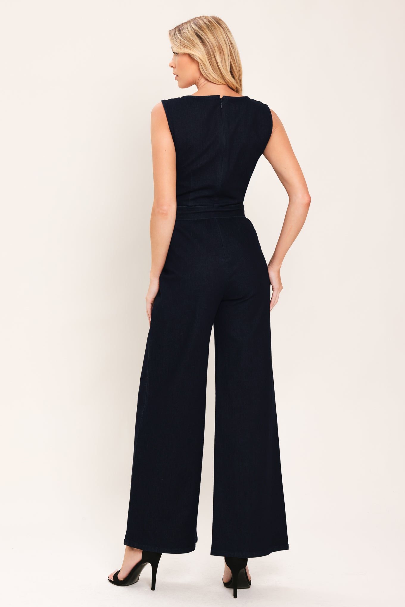 Woman wearing a dark indigo denim jumpsuit featuring sweetheart neckline, sleeveless, self belt, wide leg and back zipper closure | Flying Tomato