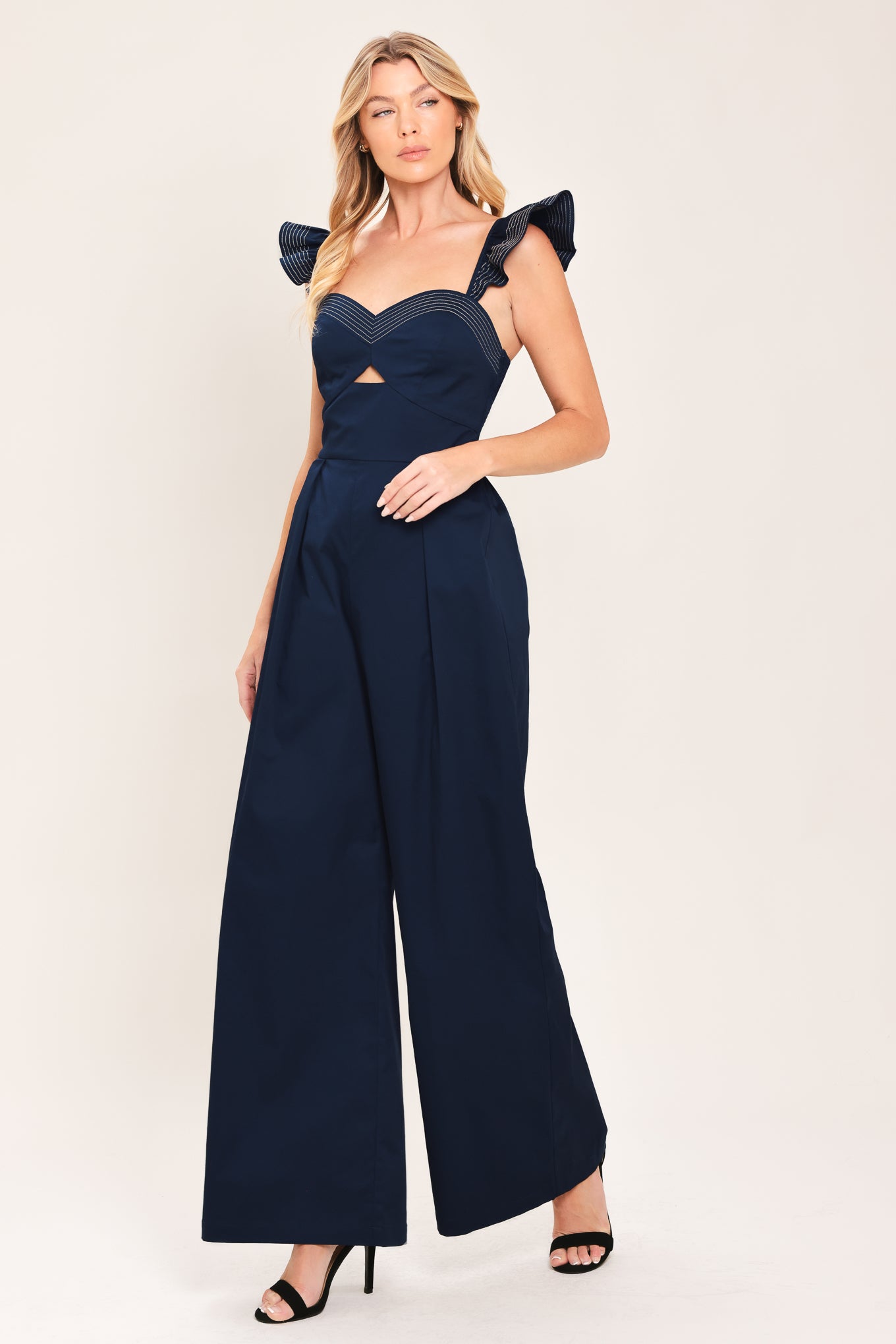 A woman wearing a navy blue wide-leg jumpsuit with ruffled shoulder details and a sweetheart neckline with a small chest cutout | Flying Tomato