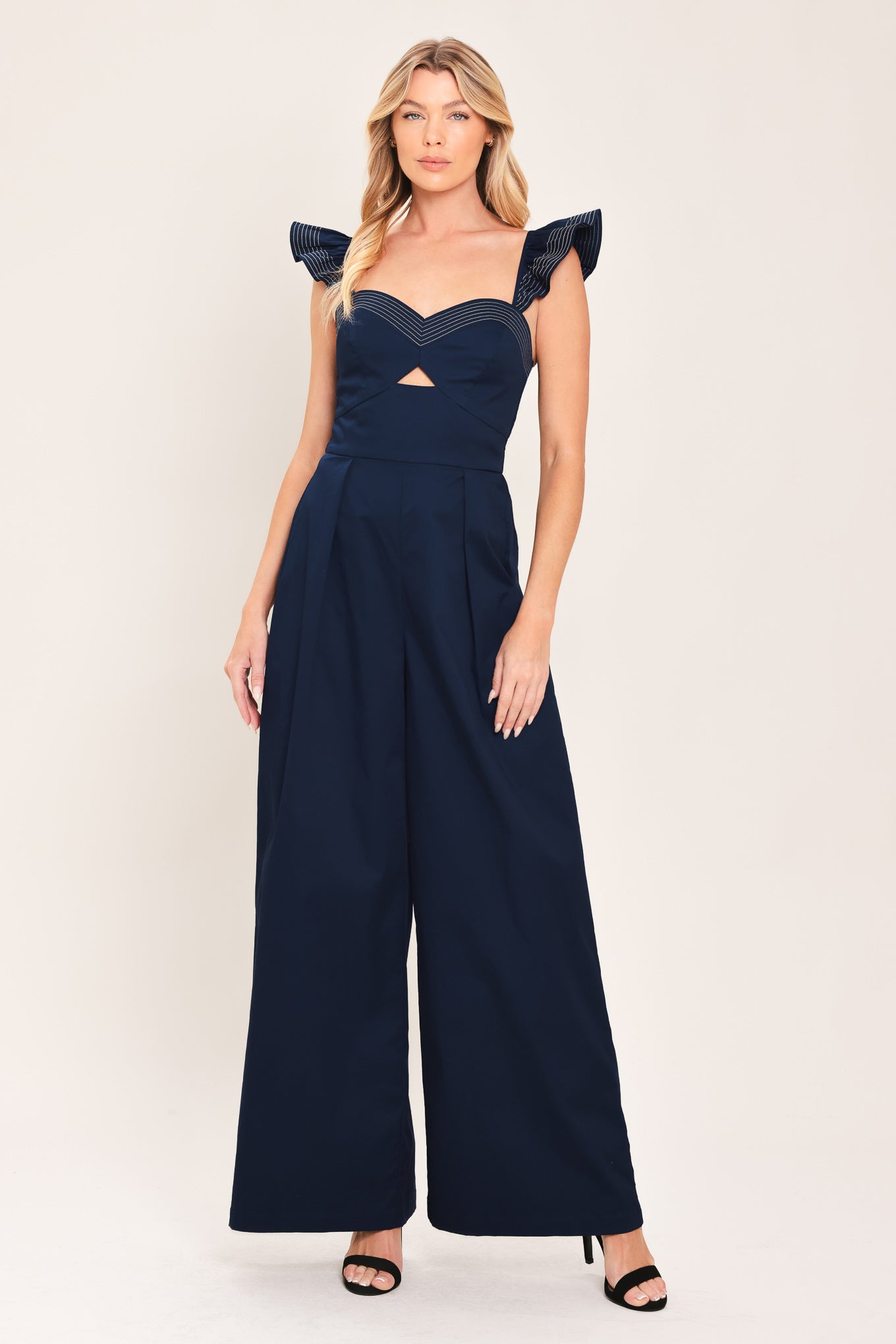 A woman wearing a navy blue wide-leg jumpsuit with ruffled shoulder details and a sweetheart neckline with a small chest cutout | Flying Tomato