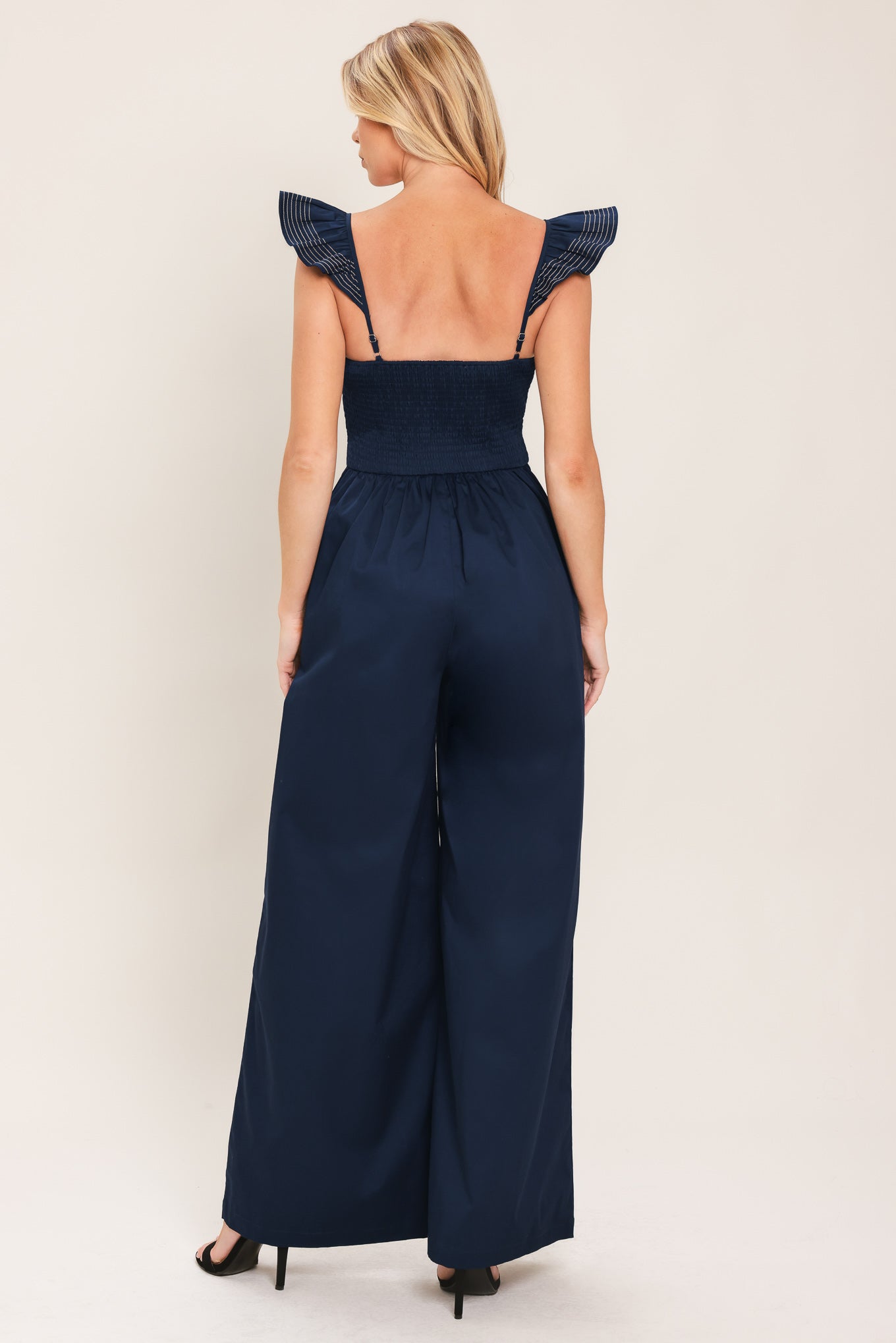 A woman wearing a navy blue wide-leg jumpsuit with ruffled shoulder details and a sweetheart neckline with a small chest cutout | Flying Tomato