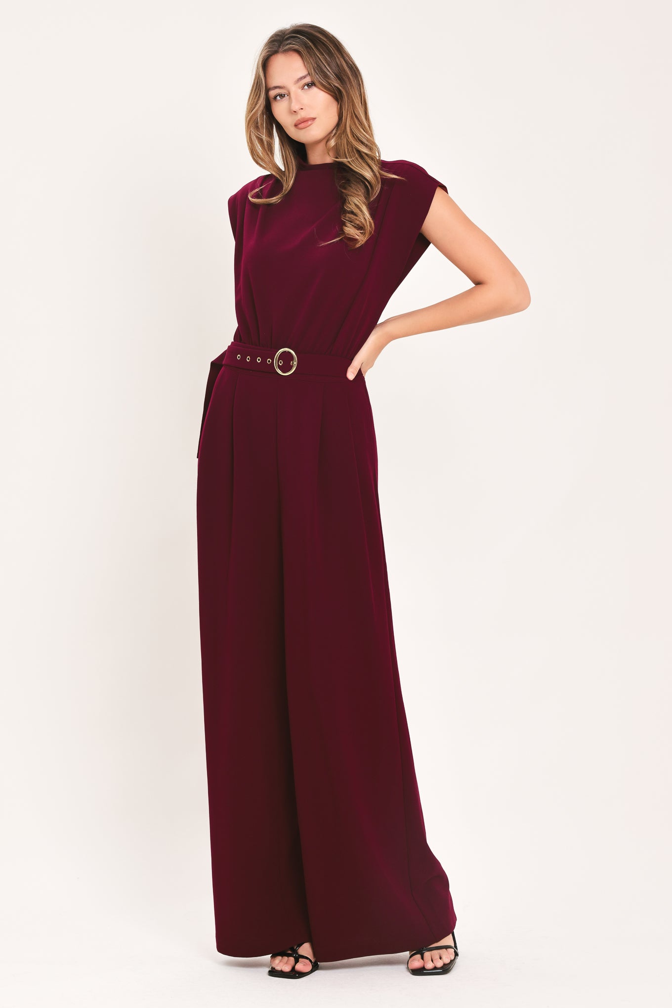 A woman modeling a sleeveless, high-neck, wide-leg jumpsuit in deep burgundy. The jumpsuit features a matching belt with a gold-tone buckle at the waist | Flying Tomato