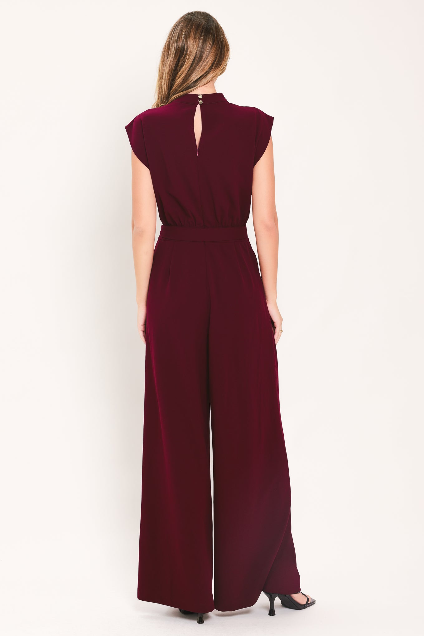 A woman modeling a sleeveless, high-neck, wide-leg jumpsuit in deep burgundy. The jumpsuit features a matching belt with a gold-tone buckle at the waist | Flying Tomato