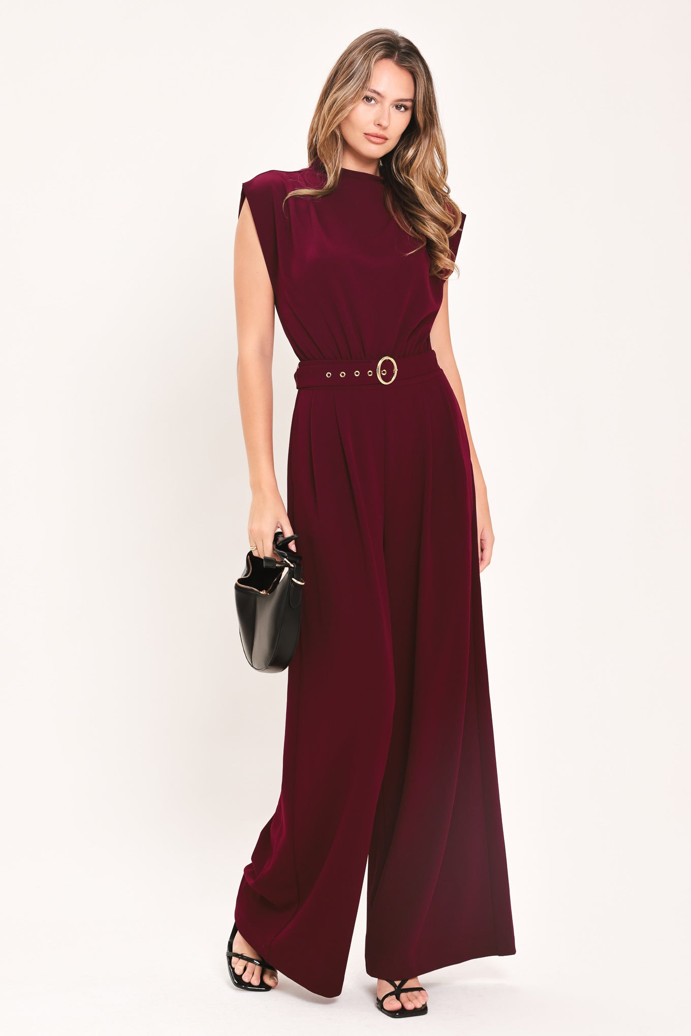 A woman modeling a sleeveless, high-neck, wide-leg jumpsuit in deep burgundy. The jumpsuit features a matching belt with a gold-tone buckle at the waist | Flying Tomato