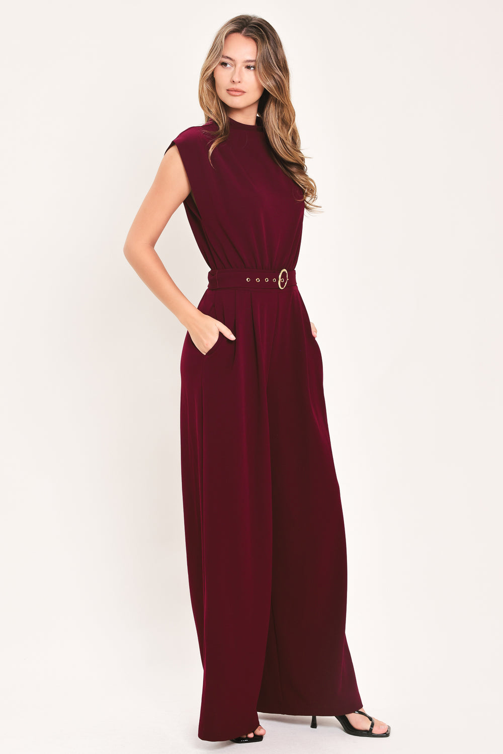 A woman modeling a sleeveless, high-neck, wide-leg jumpsuit in deep burgundy. The jumpsuit features a matching belt with a gold-tone buckle at the waist | Flying Tomato