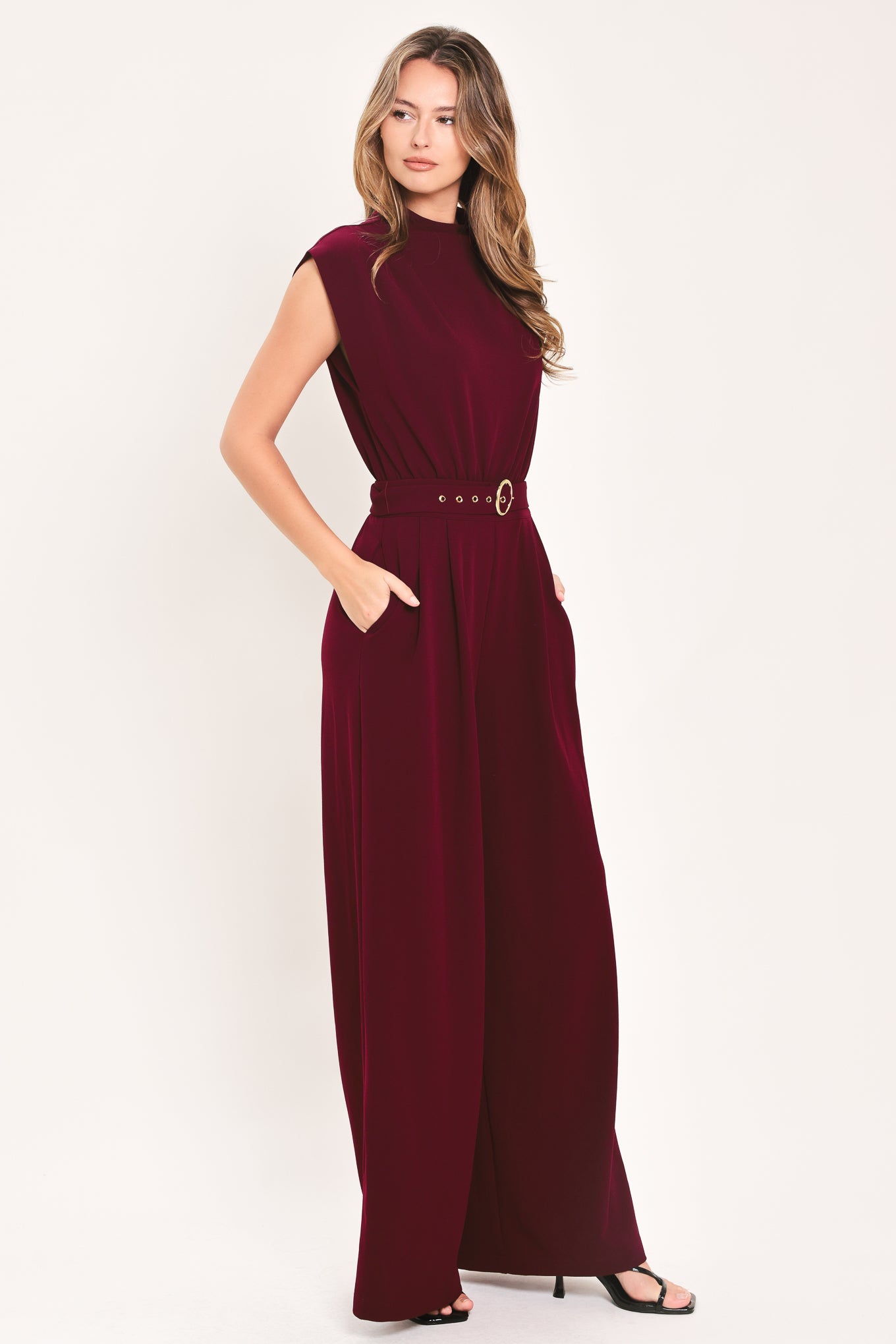 A woman modeling a sleeveless, high-neck, wide-leg jumpsuit in deep burgundy. The jumpsuit features a matching belt with a gold-tone buckle at the waist | Flying Tomato