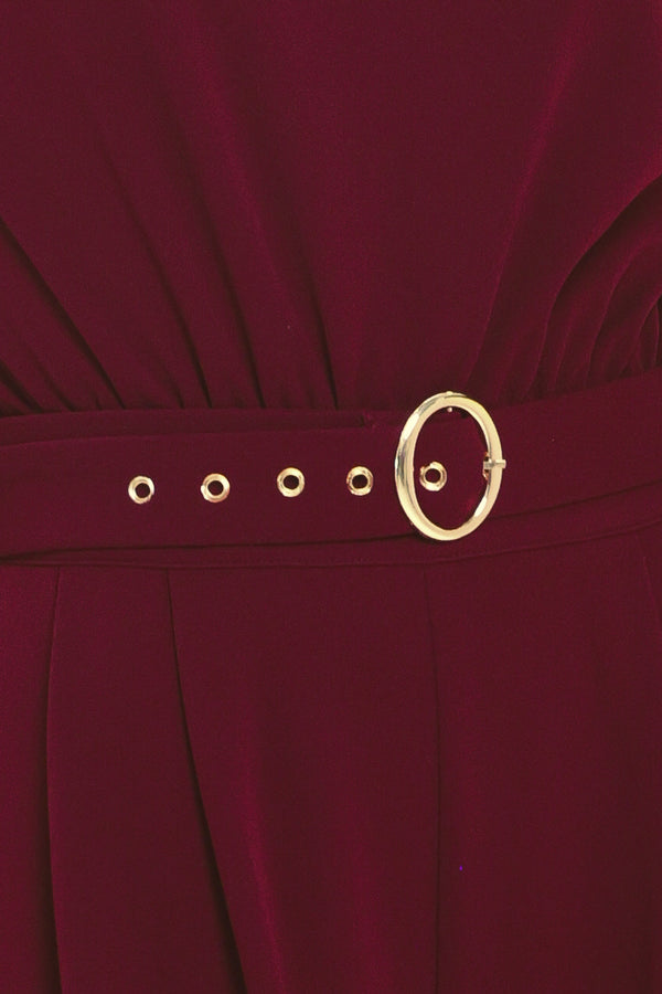 A woman modeling a sleeveless, high-neck, wide-leg jumpsuit in deep burgundy. The jumpsuit features a matching belt with a gold-tone buckle at the waist | Flying Tomato