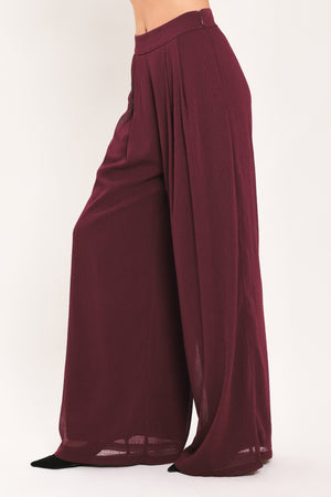 A pair of wide-leg, high-waisted trousers in a deep burgundy or maroon fabric, shown on a model | Flying Tomato