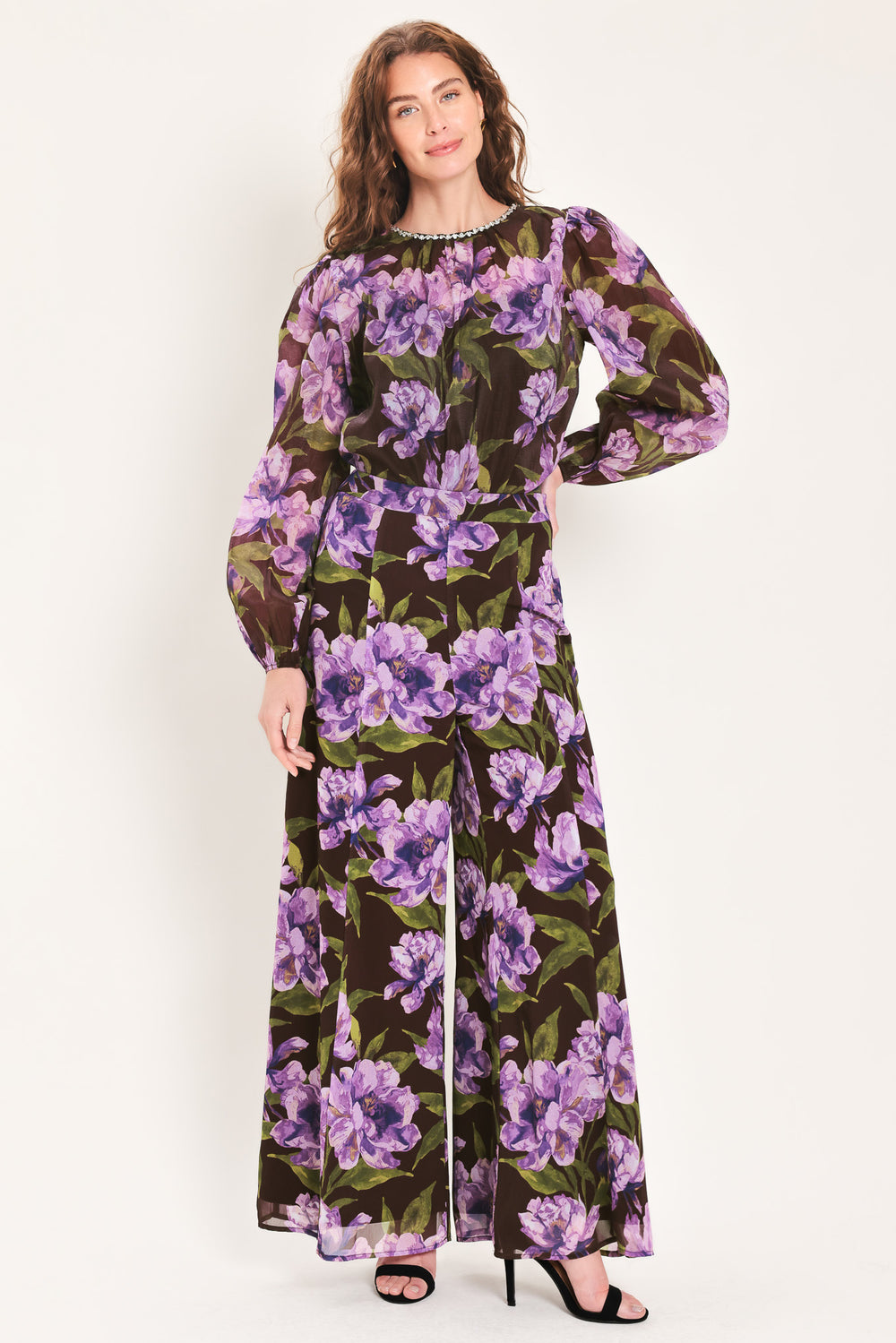 A model is wearing a pair of wide-leg, high-waisted pants featuring a bold purple floral print on a dark brown background | Flying Tomato