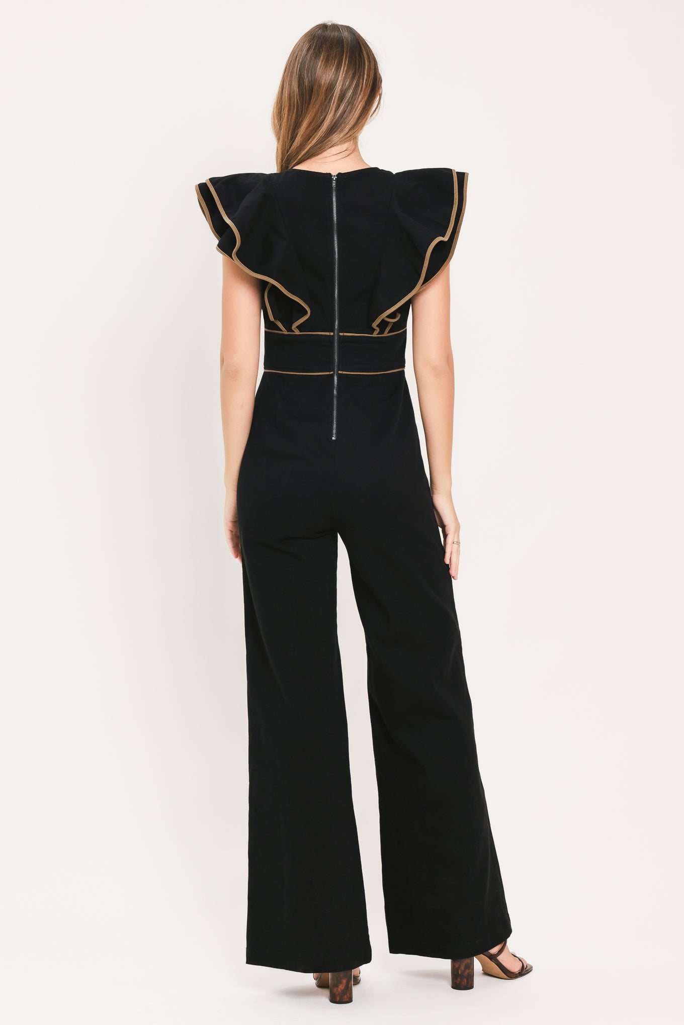 A woman modeling a black jumpsuit with dramatic ruffled cap sleeves, a deep V-neckline, and beige contrast piping details | Flying Tomato
