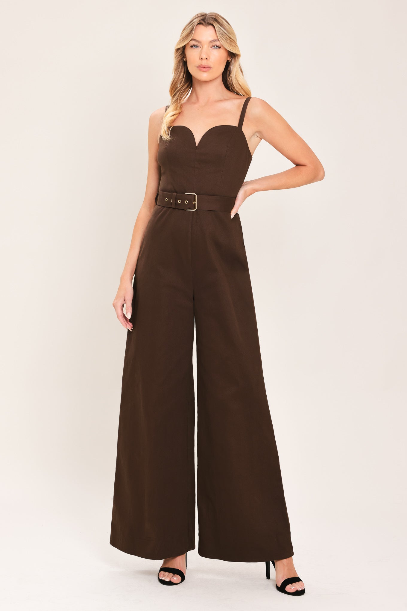 Woman wearing a brown solid woven jumpsuit featuring sweetheart neckline, straps, self belt, wide leg and back zipper closure | Flying Tomato