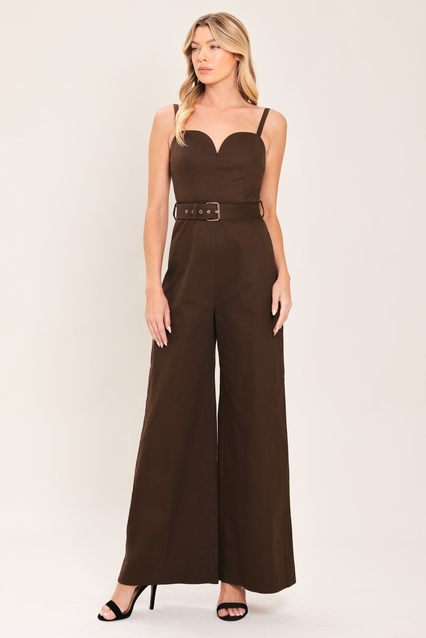 Woman wearing a brown solid woven jumpsuit featuring sweetheart neckline, straps, self belt, wide leg and back zipper closure | Flying Tomato