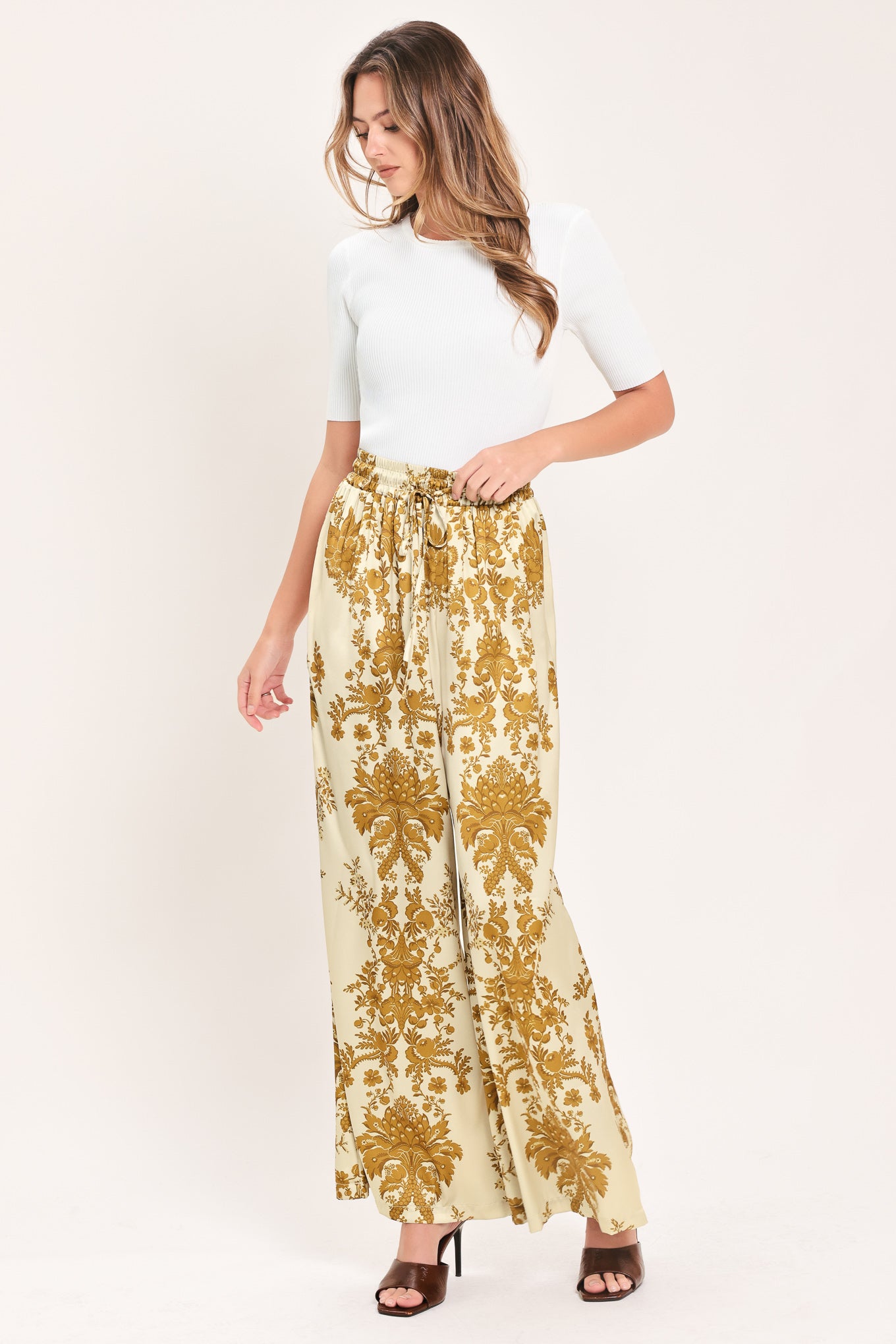 A woman wearing high-waisted, wide-leg pants with a beige base and an ornate gold damask pattern | Flying Tomato