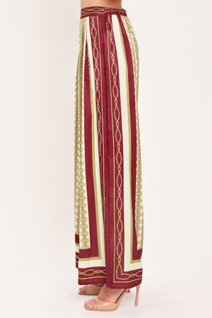 A woman wearing a pair of wide-leg pants with a beige and gold geometric pattern, featuring deep burgundy side panels and a matching patterned hem | Flying Tomato