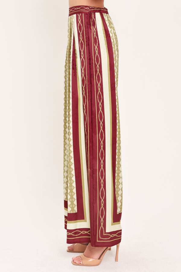 A woman wearing a pair of wide-leg pants with a beige and gold geometric pattern, featuring deep burgundy side panels and a matching patterned hem | Flying Tomato