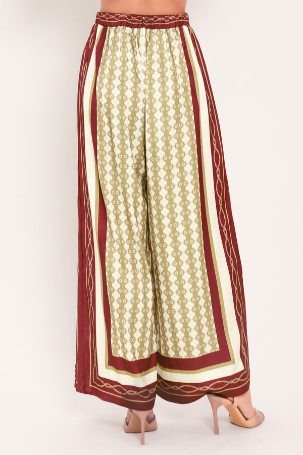 A woman wearing a pair of wide-leg pants with a beige and gold geometric pattern, featuring deep burgundy side panels and a matching patterned hem | Flying Tomato