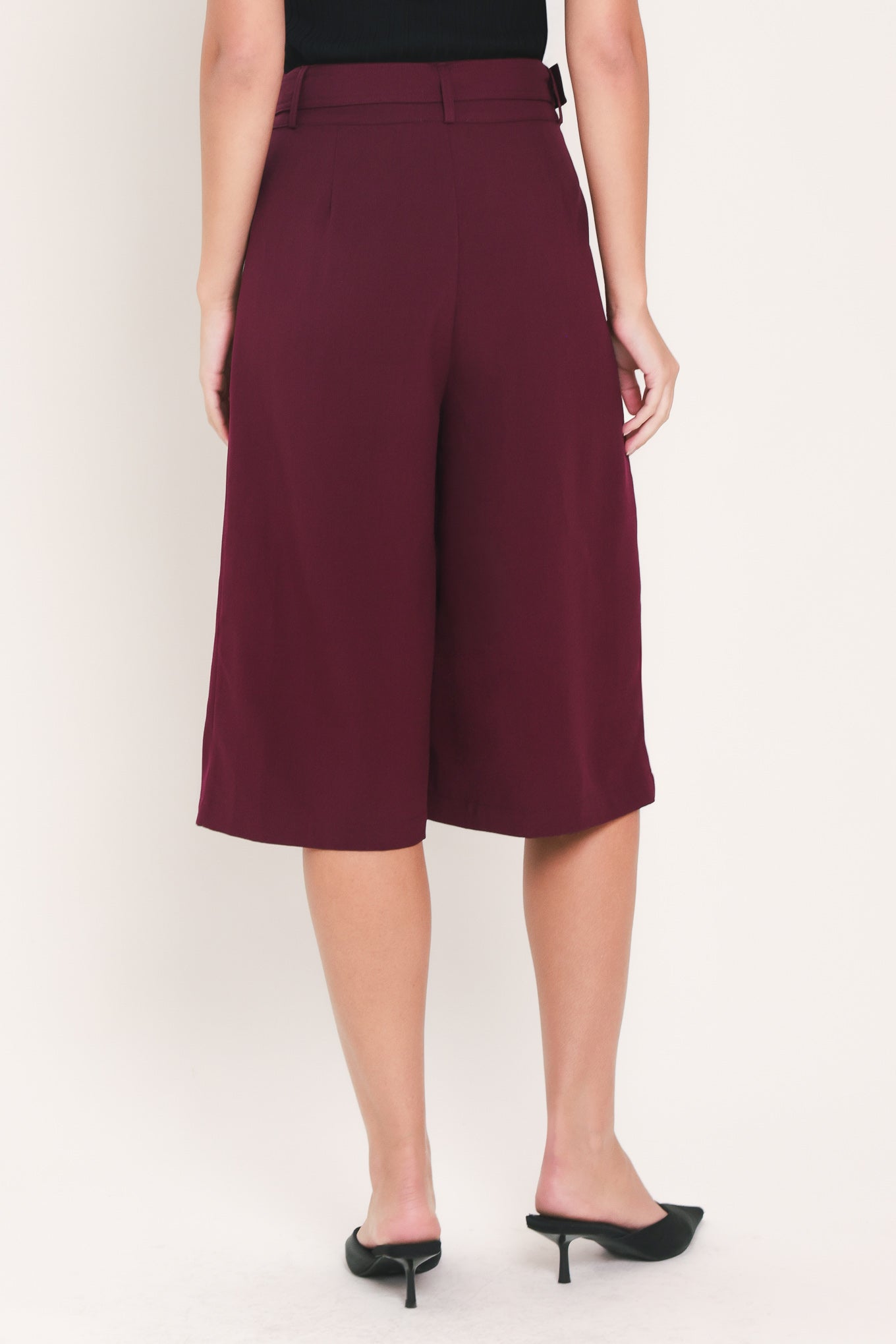 A model wearing high-waisted, wide-leg cropped trousers in deep burgundy with pleats and a matching belt | Flying Tomato