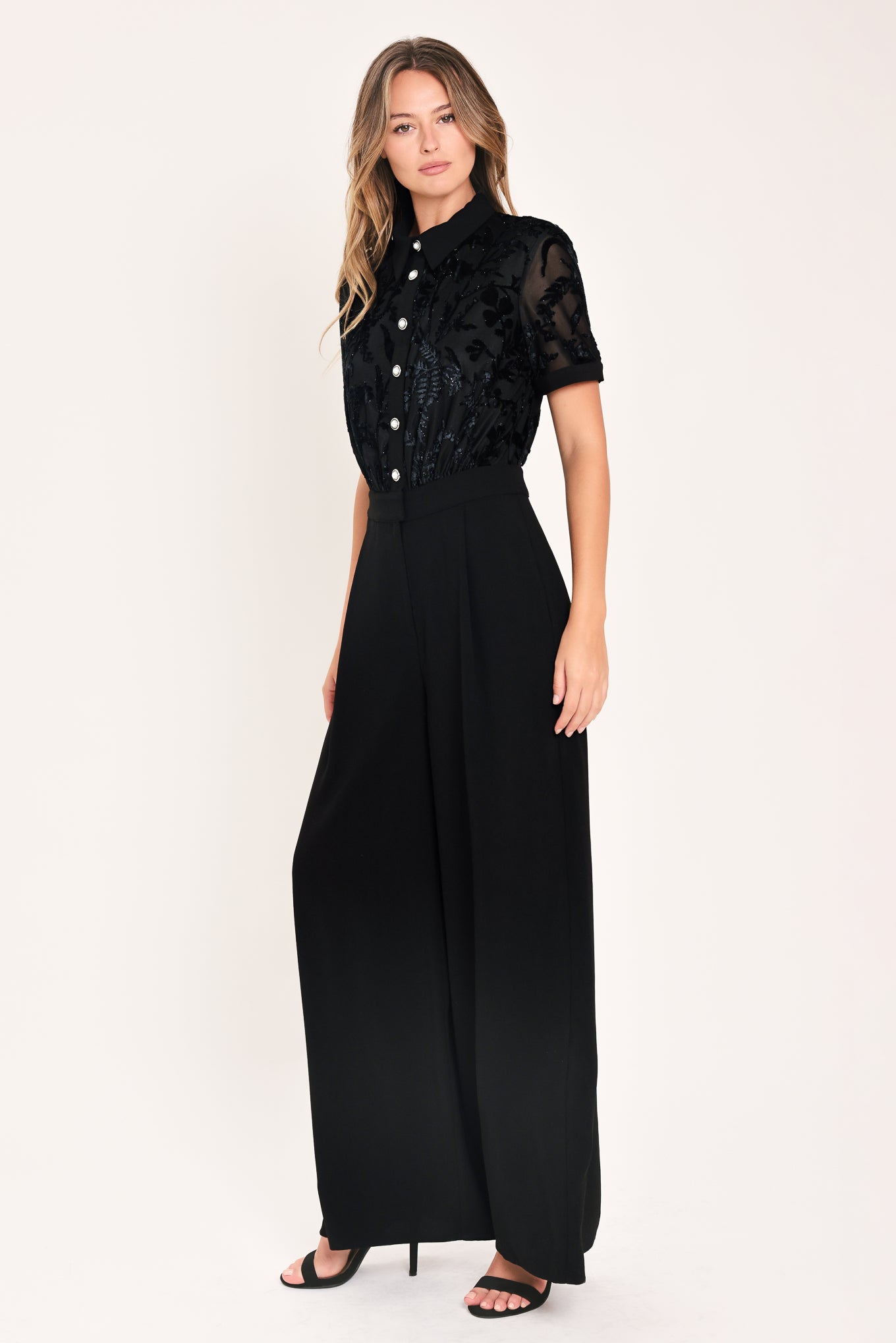 A woman modeling a black short-sleeve jumpsuit with a ruffled button-front bodice and wide-leg pants | Flying Tomato