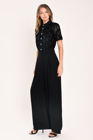 A woman modeling a black short-sleeve jumpsuit with a ruffled button-front bodice and wide-leg pants | Flying Tomato