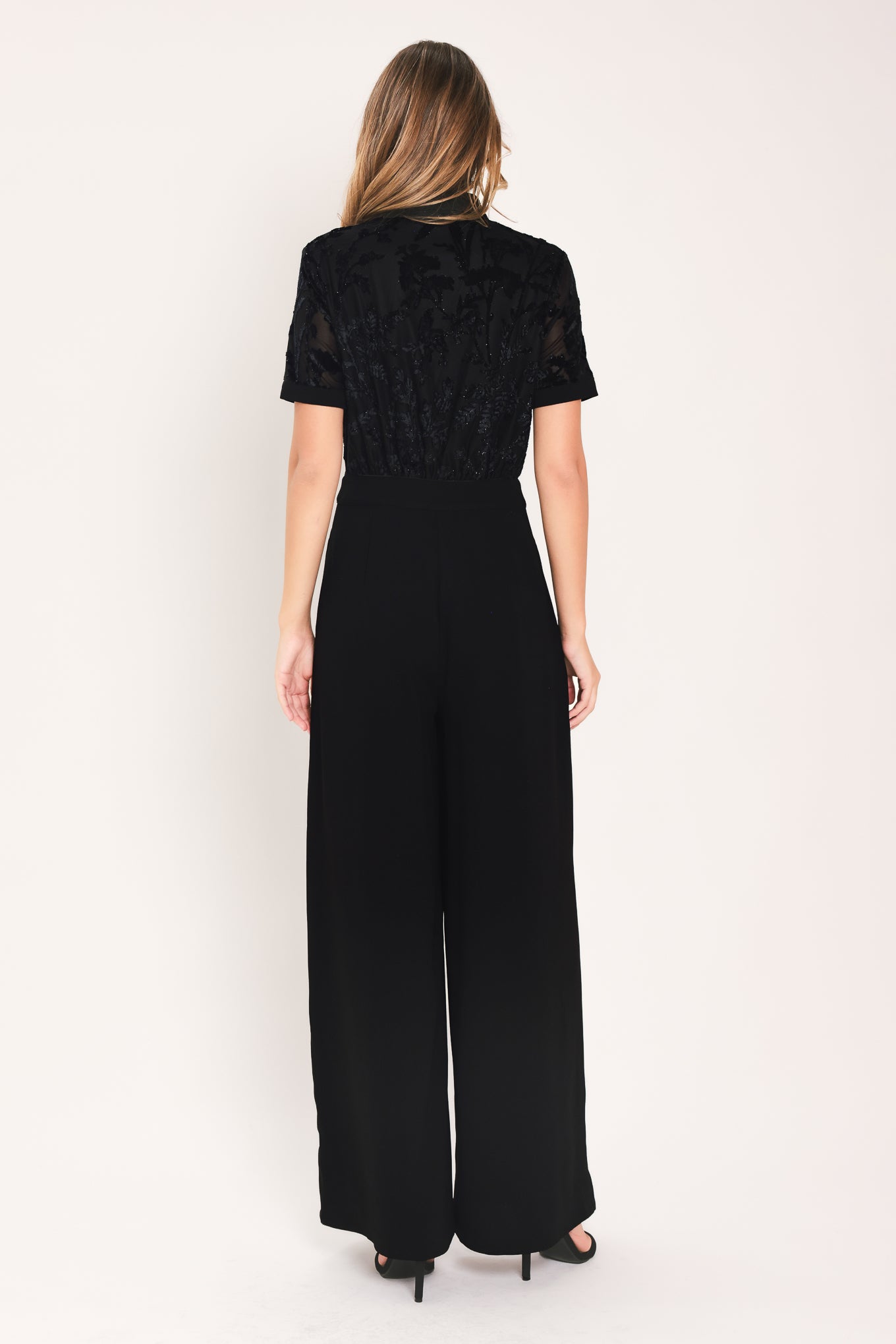 A woman modeling a black short-sleeve jumpsuit with a ruffled button-front bodice and wide-leg pants | Flying Tomato