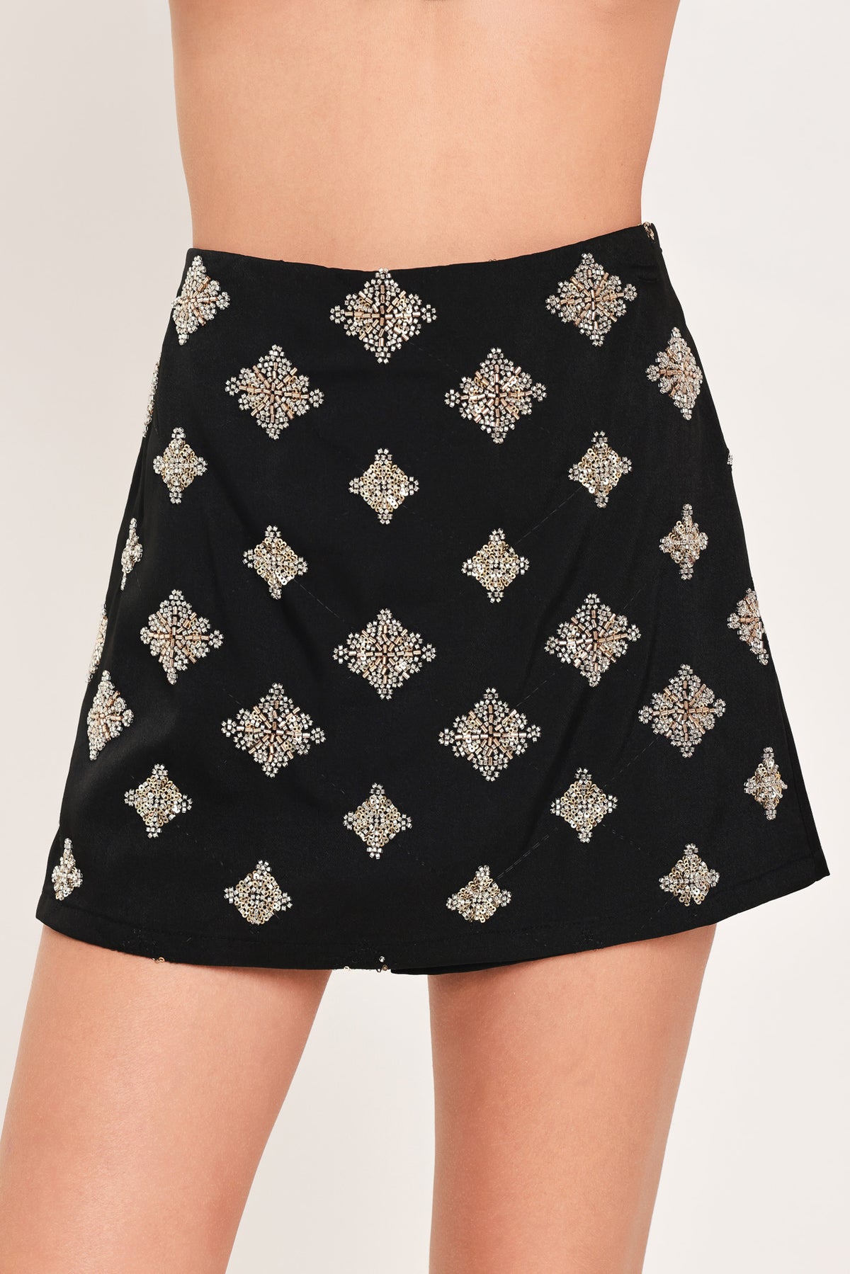A black A-line mini skort with an allover pattern of metallic gold and silver rhinestone medallions, shown on a model | Flying Tomato
