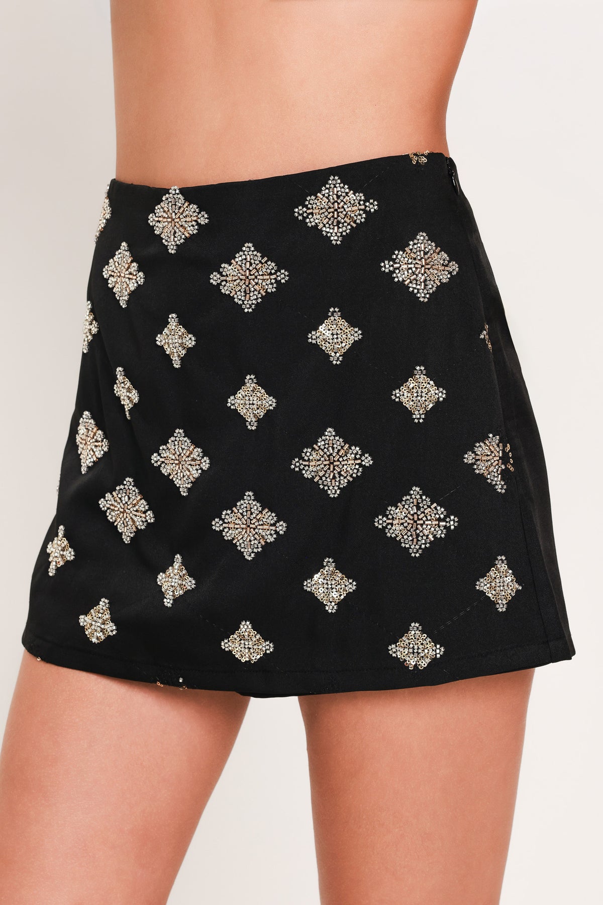 A black A-line mini skort with an allover pattern of metallic gold and silver rhinestone medallions, shown on a model | Flying Tomato