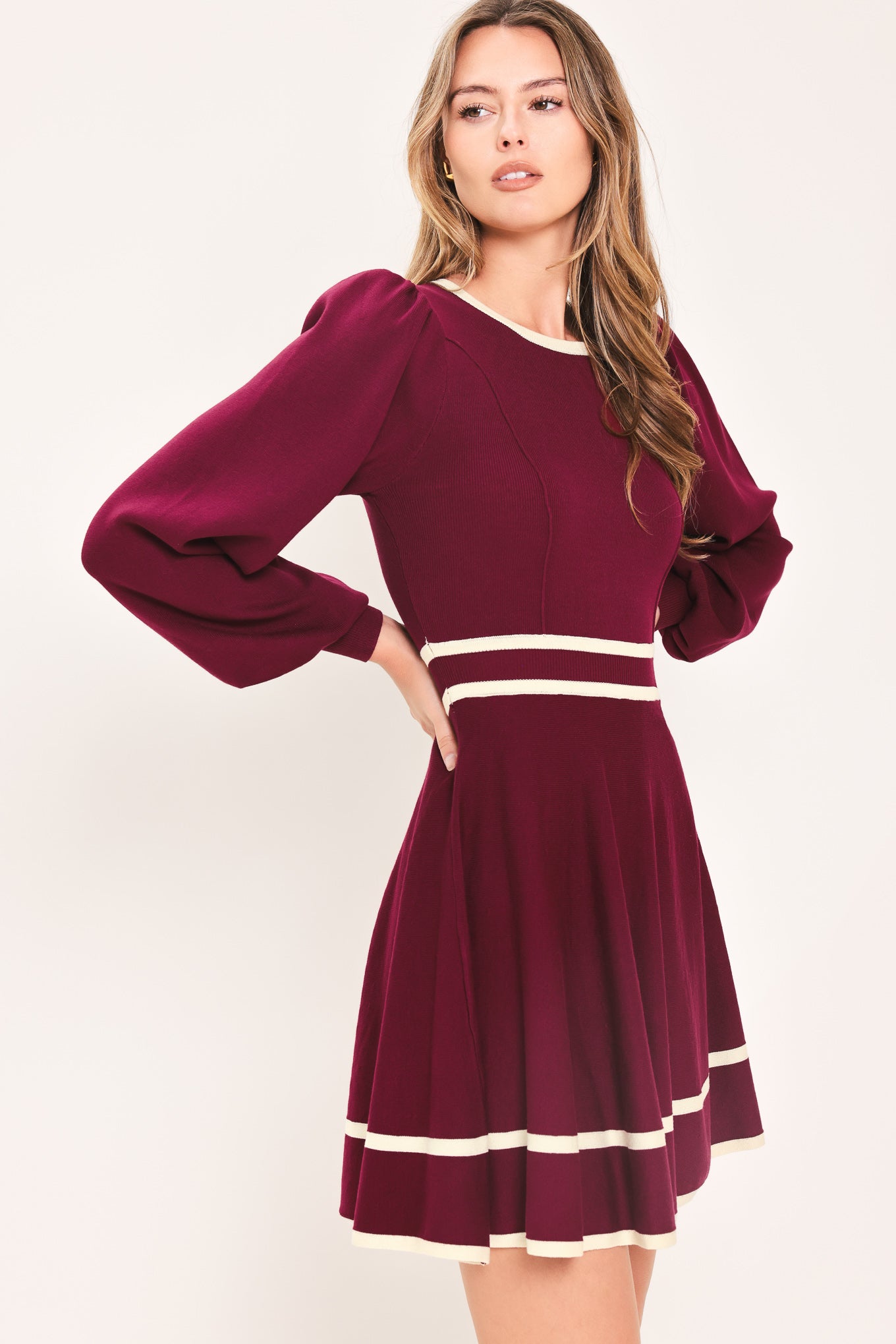 A woman wearing a long-sleeve skater dress in deep wine with cream stripe accents at the waist and hem | Flying Tomato