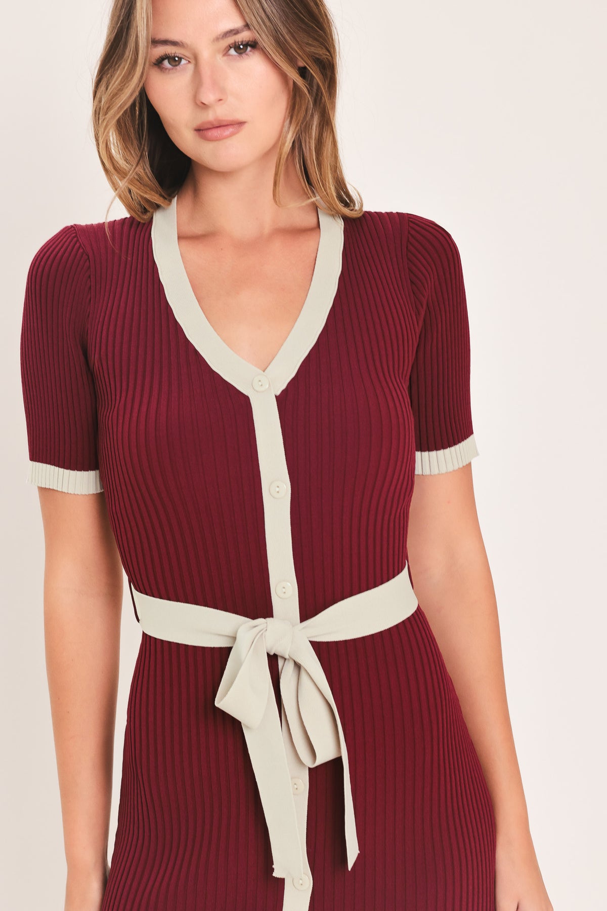 A woman models a short-sleeve ribbed knit midi dress in deep burgundy with cream trim details, including the collar, front placket, and matching tie belt | Flying Tomato