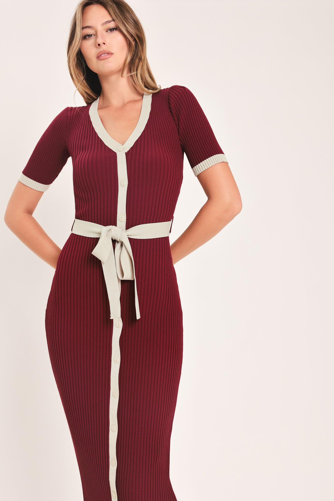 A woman models a short-sleeve ribbed knit midi dress in deep burgundy with cream trim details, including the collar, front placket, and matching tie belt | Flying Tomato