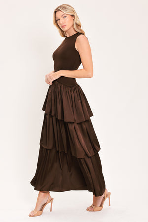 A woman modeling a sleeveless, high-neck, tiered skirt maxi dress in a deep chocolate brown color with an asymmetrical drop waist | Flying Tomato
