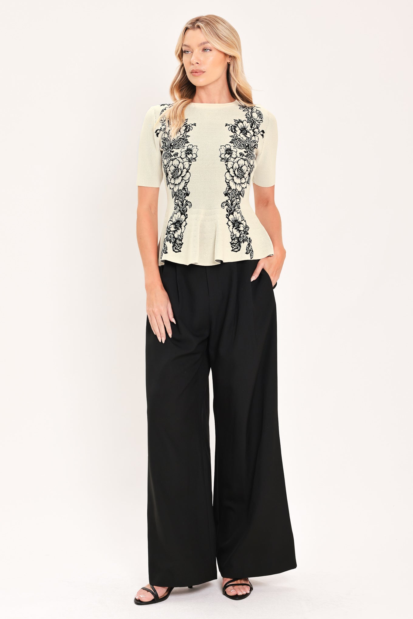 A woman modeling a cream-colored short-sleeve peplum top with black floral embroidery down the front | Flying Tomato