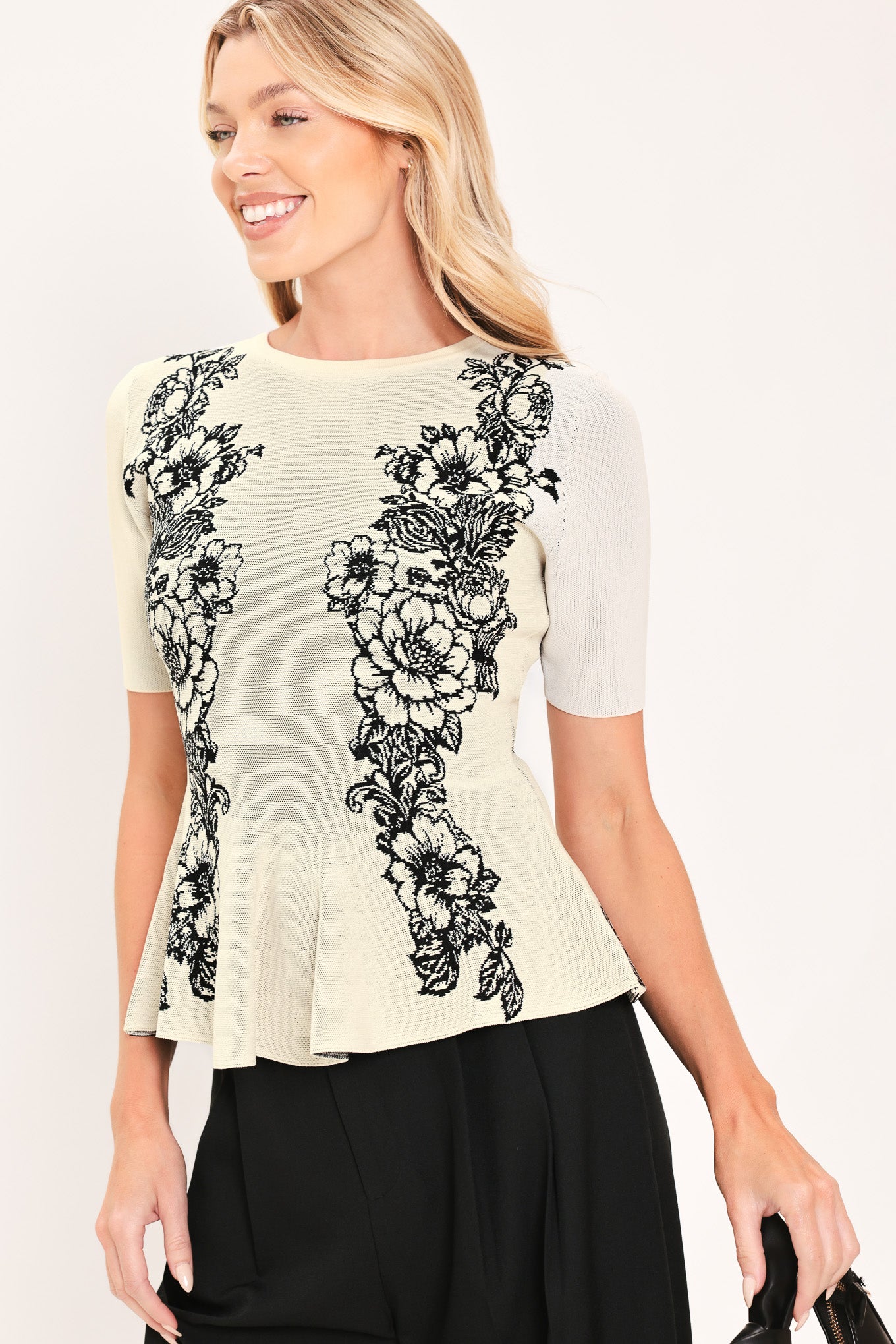 A woman modeling a cream-colored short-sleeve peplum top with black floral embroidery down the front | Flying Tomato
