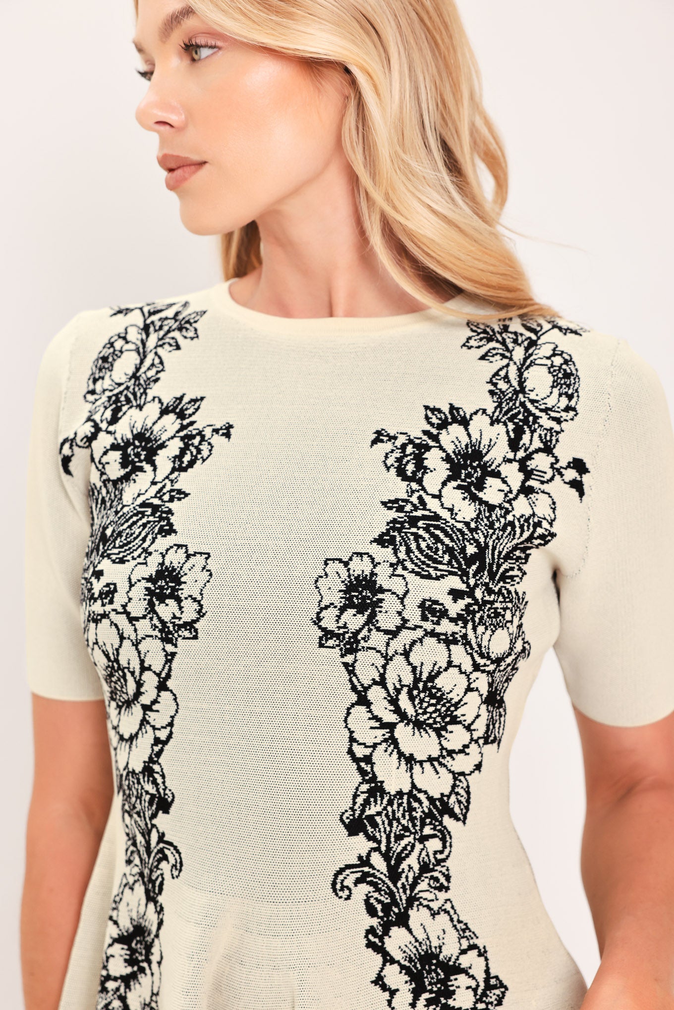 A woman modeling a cream-colored short-sleeve peplum top with black floral embroidery down the front | Flying Tomato