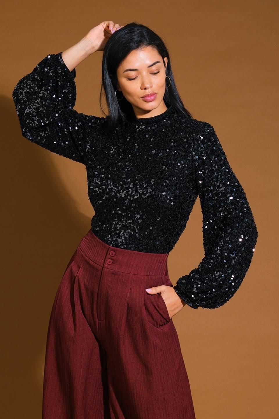 A woman modeling a black sequined long-sleeve mock neck top paired with burgundy pleated trousers, set against a plain off-white studio background.