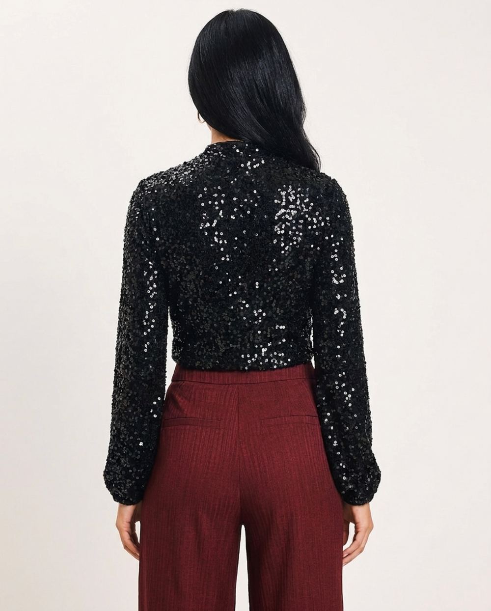 A woman modeling a black sequined long-sleeve mock neck top paired with burgundy pleated trousers, set against a plain off-white studio background.