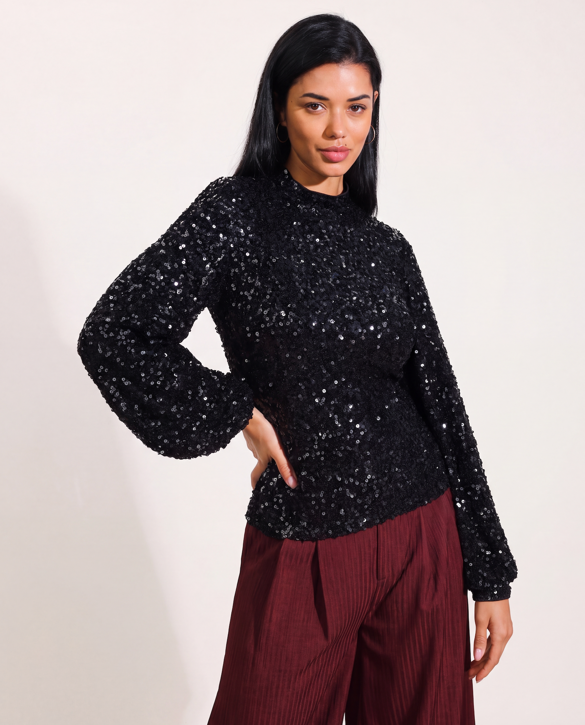 A woman modeling a black sequined long-sleeve mock neck top paired with burgundy pleated trousers, set against a plain off-white studio background.