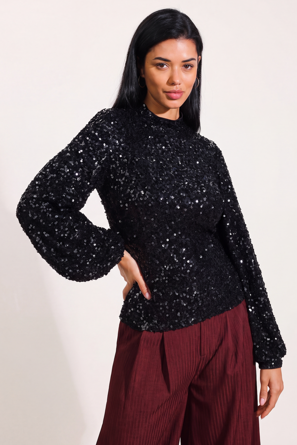 A woman modeling a black sequined long-sleeve mock neck top paired with burgundy pleated trousers, set against a plain off-white studio background.