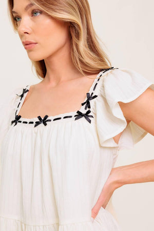 A solid woven top featuring square neckline, short flutter sleeve, ribbon trim and ruffled hemline.
