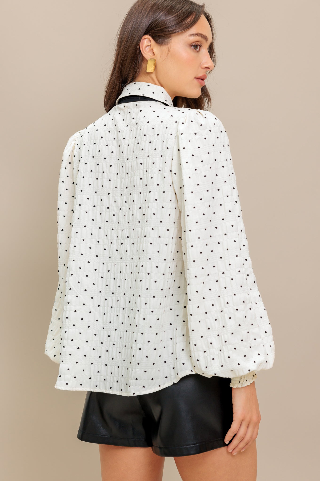 A heart printed woven top featuring collar, contrasting bow tie, long sleeve with smocked cuff and front button closure