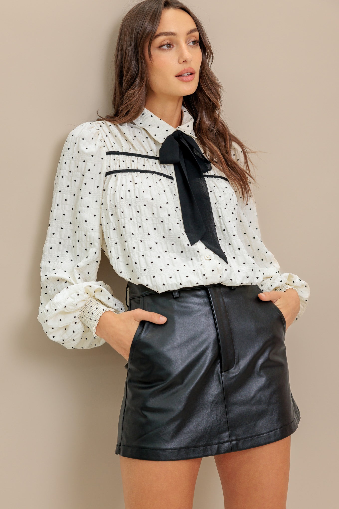 A heart printed woven top featuring collar, contrasting bow tie, long sleeve with smocked cuff and front button closure