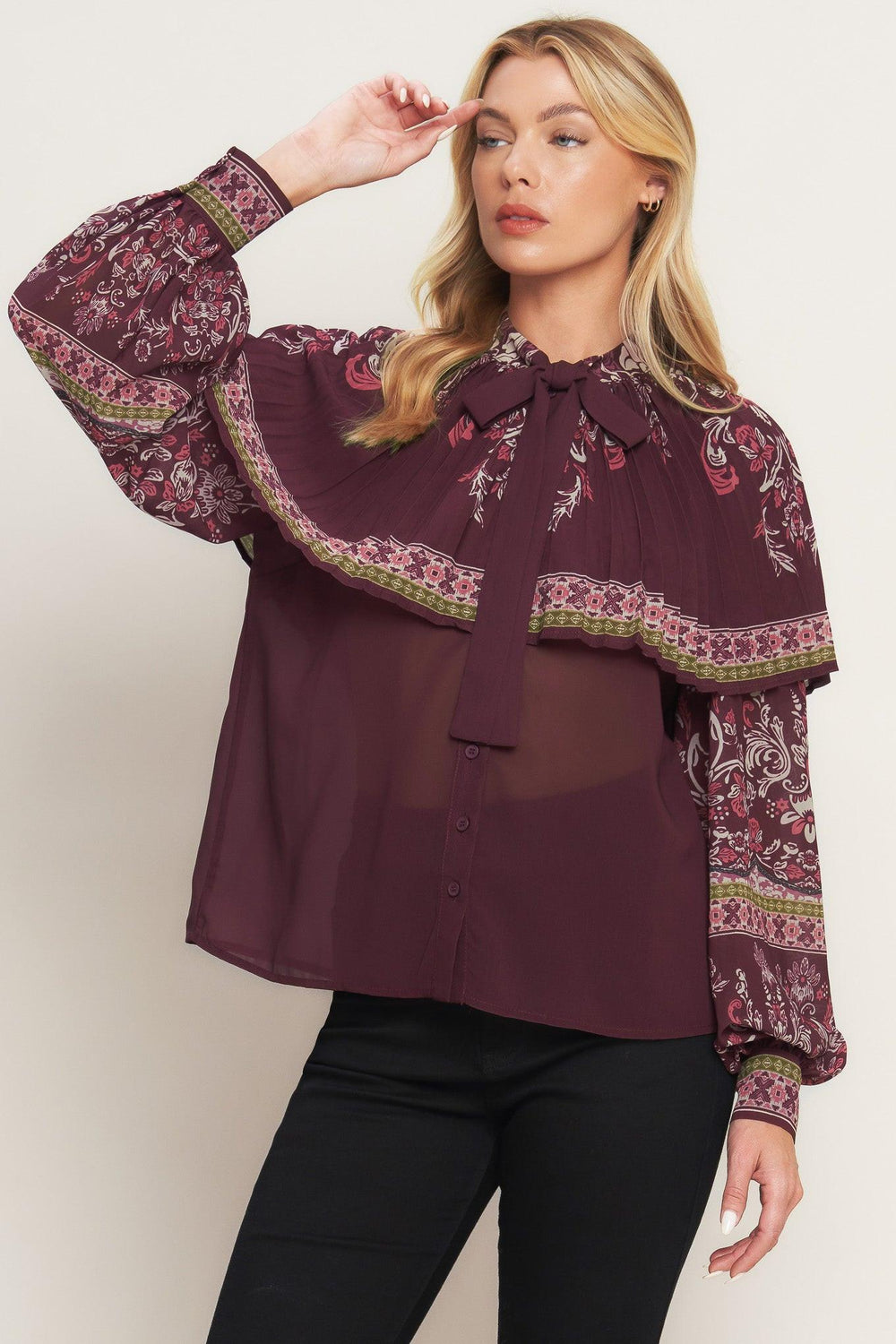 A printed woven top featuring pleated capelet, front bow tie, button down and long sleeve with cuff. 