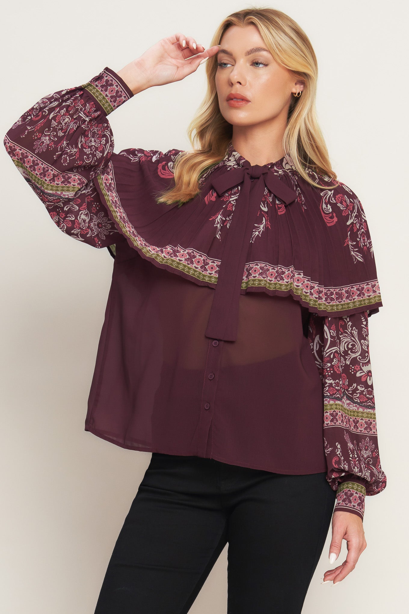 A printed woven top featuring pleated capelet, front bow tie, button down and long sleeve with cuff. 