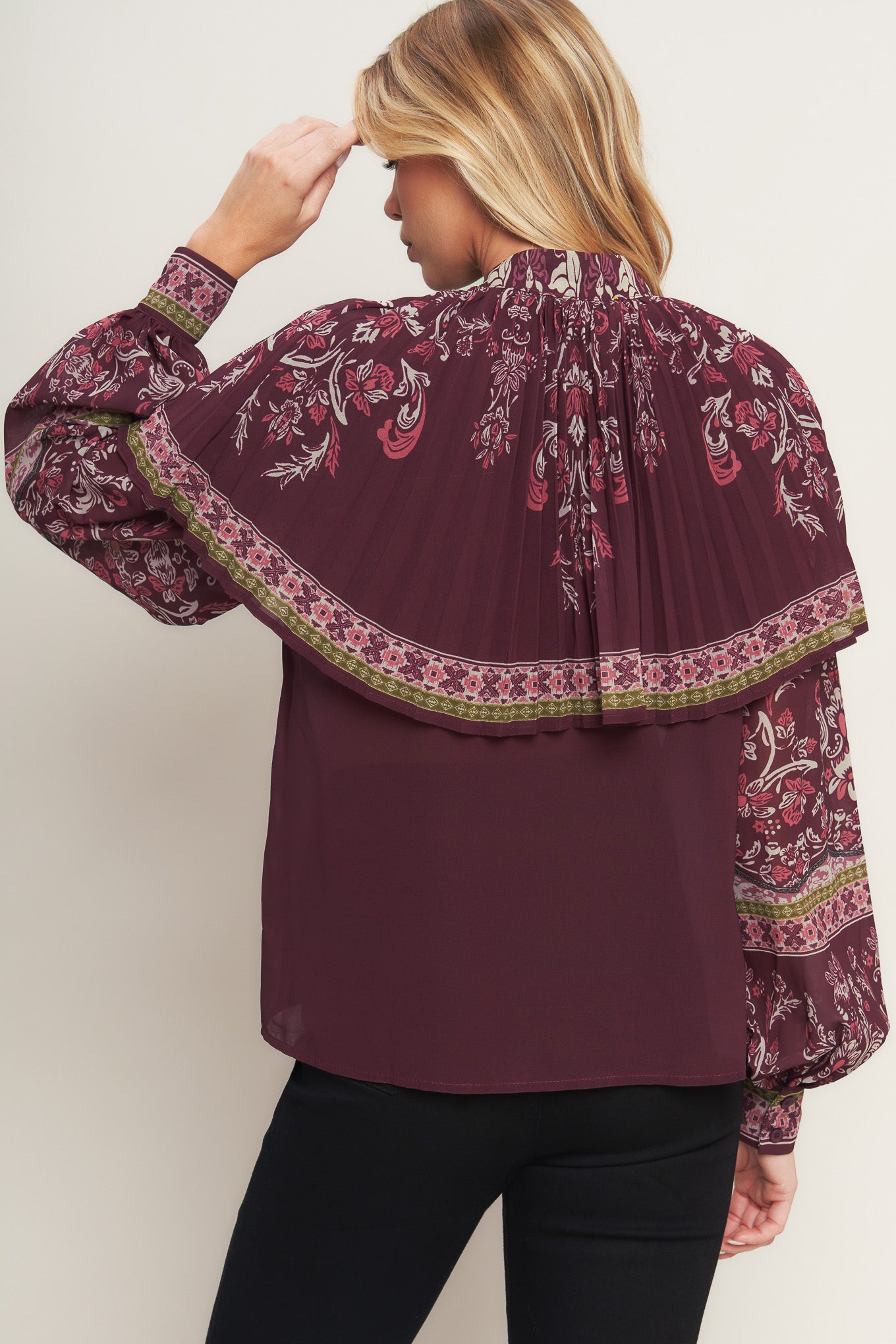 A printed woven top featuring pleated capelet, front bow tie, button down and long sleeve with cuff. 