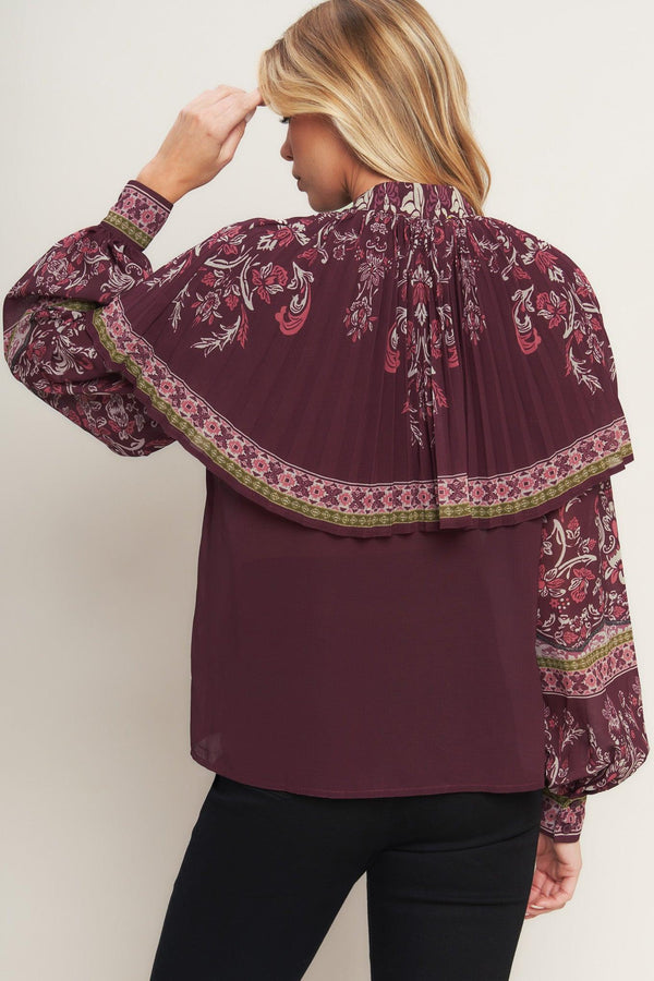 A printed woven top featuring pleated capelet, front bow tie, button down and long sleeve with cuff. 