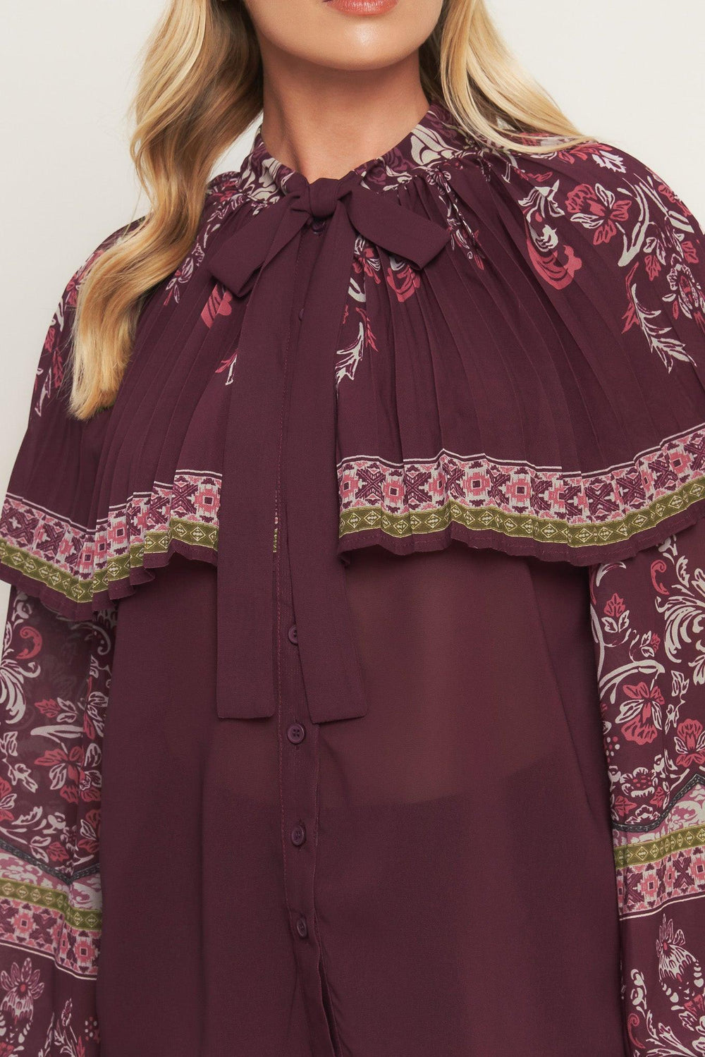 A printed woven top featuring pleated capelet, front bow tie, button down and long sleeve with cuff. 