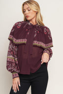 A printed woven top featuring pleated capelet, front bow tie, button down and long sleeve with cuff. 