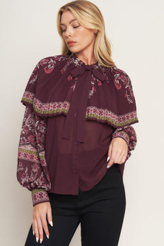 A printed woven top featuring pleated capelet, front bow tie, button down and long sleeve with cuff. 